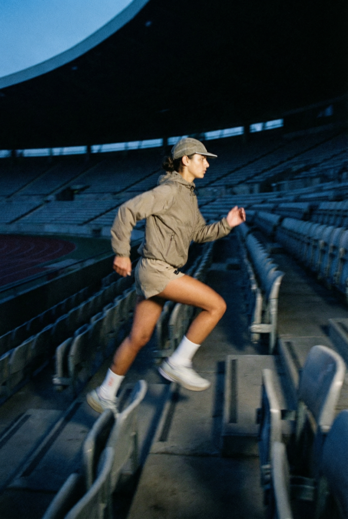AI-generated fashion editorial — female model in UNNA technical jacket running up stadium stairs at dusk, Kodachi Studio