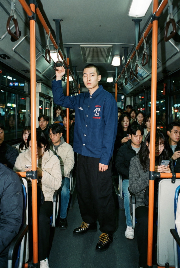 AI-generated fashion editorial — male model in SCRT coach jacket standing on crowded Korean city bus, Kodachi Studio